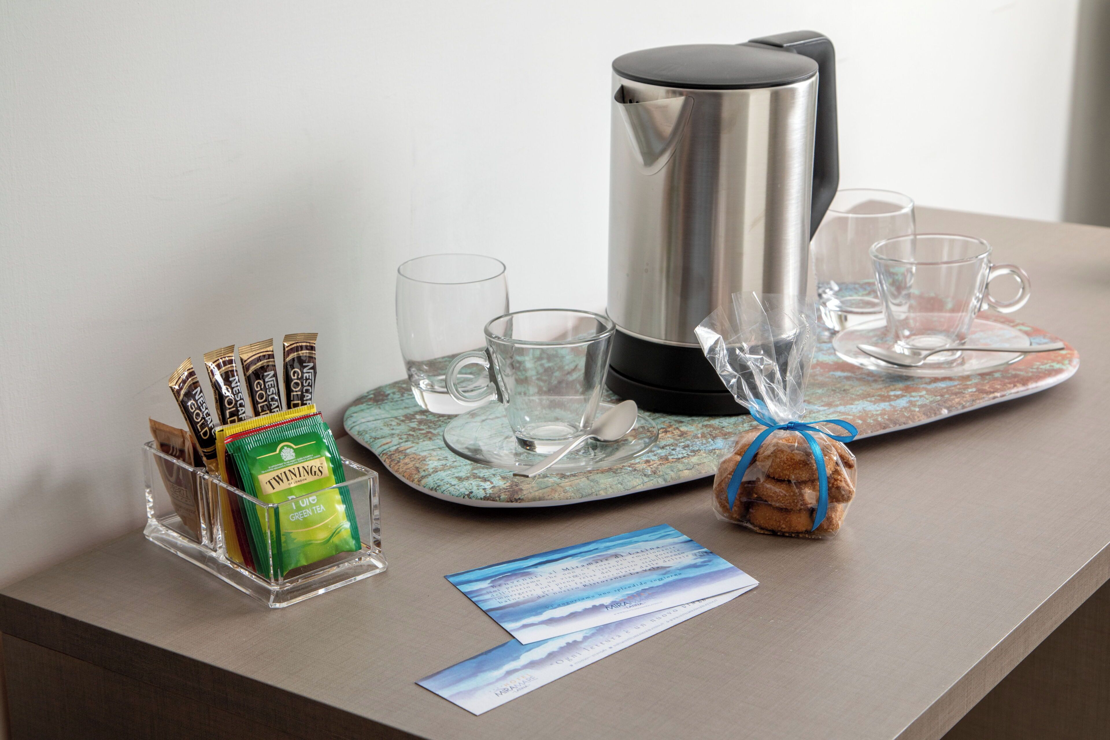 Una disposición para el desayuno con una cafetera, tazas, una bandeja, un recipiente de vidrio con bolsitas de té y un objeto pequeño envuelto sobre una mesa.