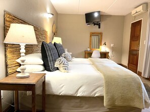 Habitación de hotel con dos camas, una mesita de noche con lámpara, un televisor montado en la pared y un cabecero de madera.