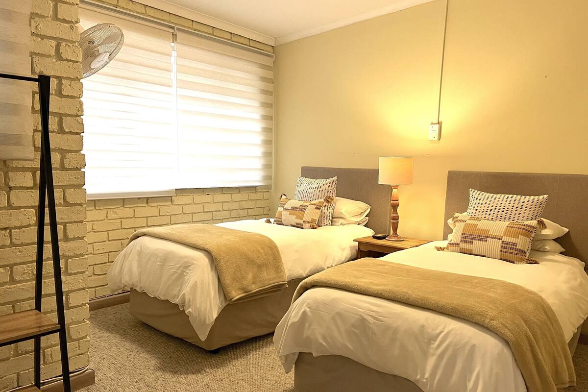 Habitación de hotel con dos camas, una pared de ladrillo, una lámpara de pie y una ventana con persianas.