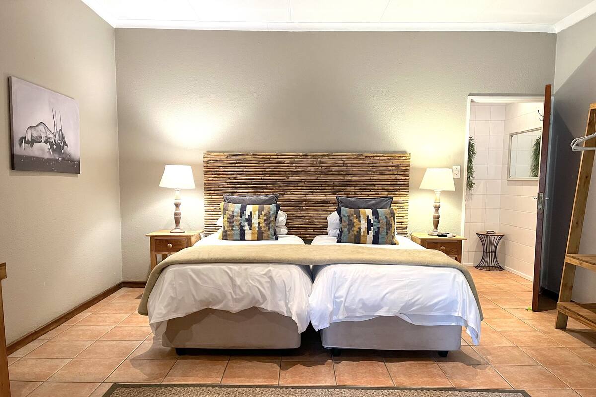 Una cama doble con cabecera texturizada, dos mesitas de noche con lámparas, un cuadro enmarcado en la pared y una pequeña mesita con una planta.