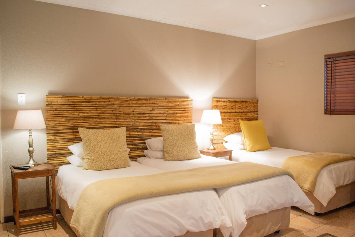 Una habitación de hotel con dos camas, cabecero de madera, lámparas de noche y una ventana con persianas.