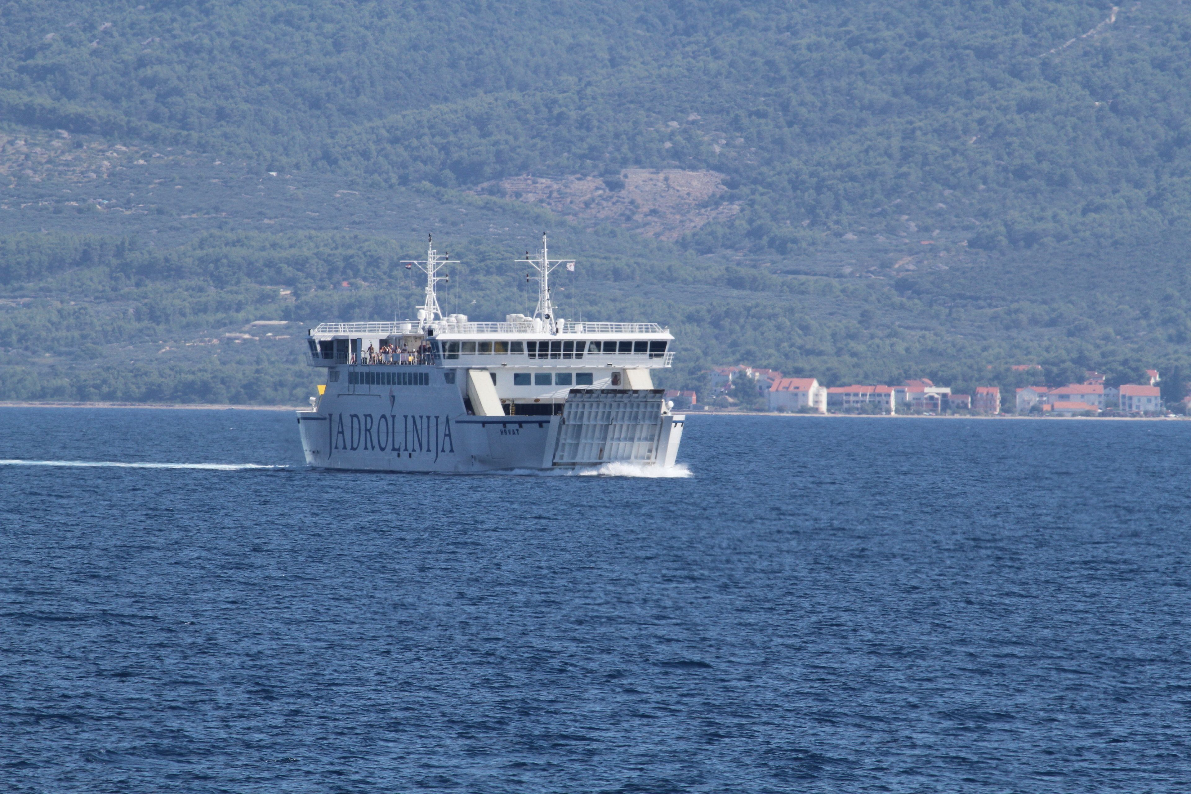 A large ferry with multiple decks and a cabin structure, sailing on a body of water with a mountainous coastline in the background.