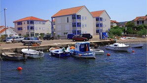 A marina with several small boats docked, a parking area with cars, and a row of buildings with red-tiled roofs. - Aparthotel Marina Gromela (Supetar)