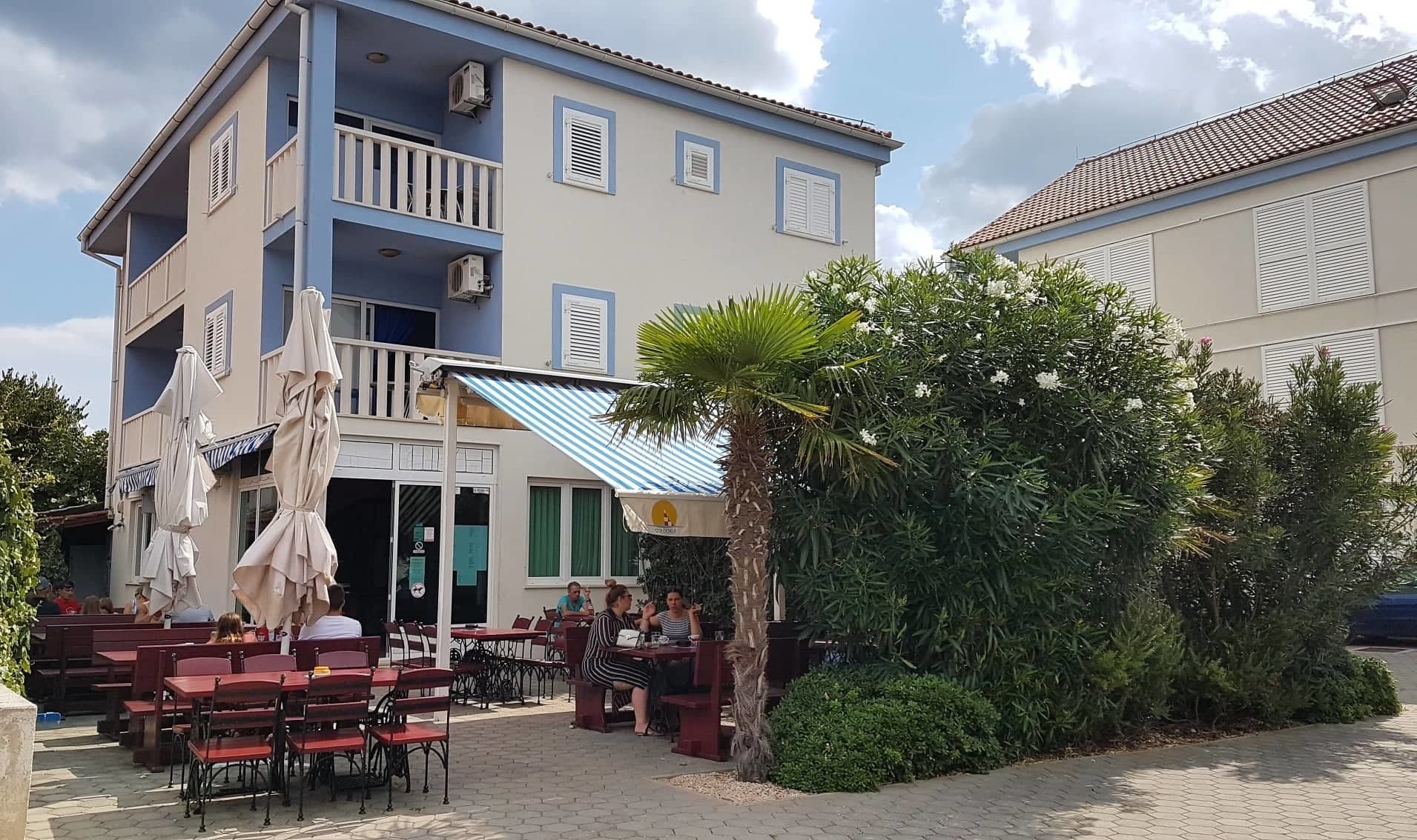 Un bâtiment à deux étages doté d’une terrasse, d’un palmier et d’une enseigne indiquant qu’il s’agit d’un restaurant.