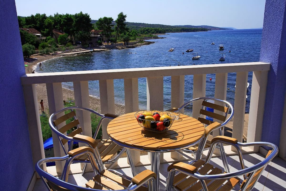 Un balcon avec une table et des chaises, un panier de fruits et une vue sur la mer, où l’on aperçoit des bateaux.