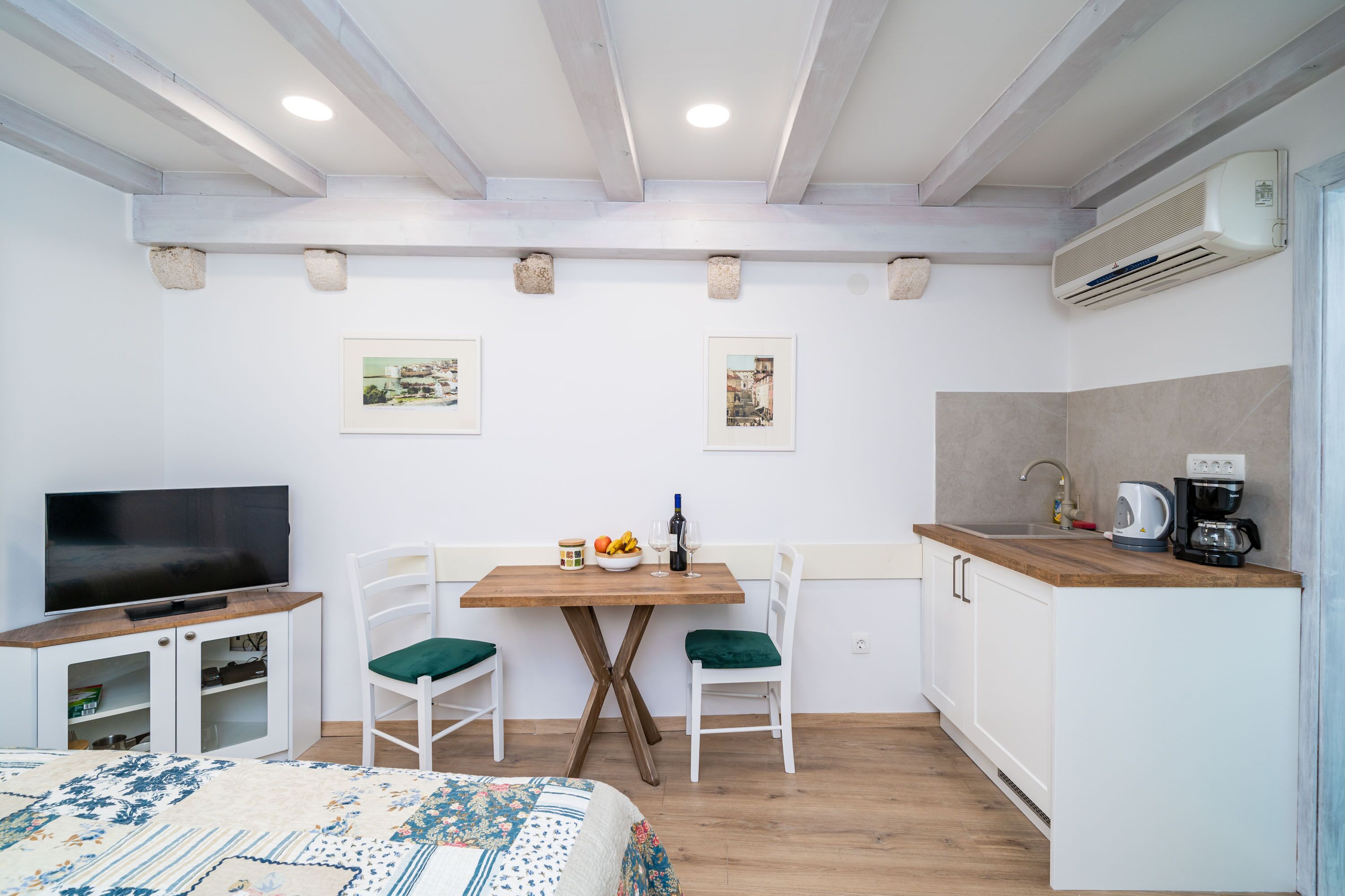 A compact room with a bed, a TV, a dining table with chairs, a kitchen area, and two framed pictures on the wall.