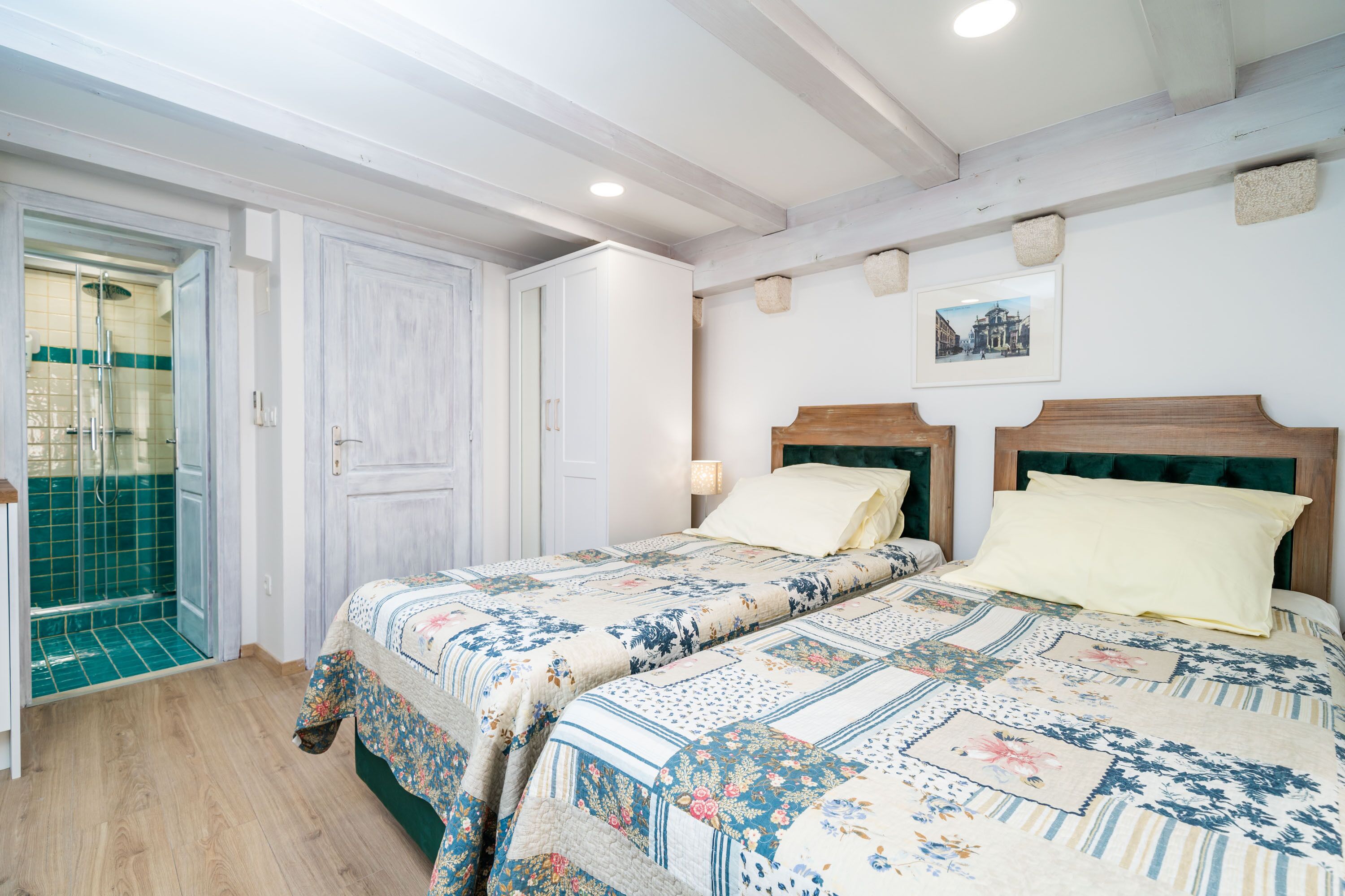 Un dormitorio con cabecera de madera, cama con ropa de cama floral, suelo de madera y baño con zona de ducha de azulejos verdes.