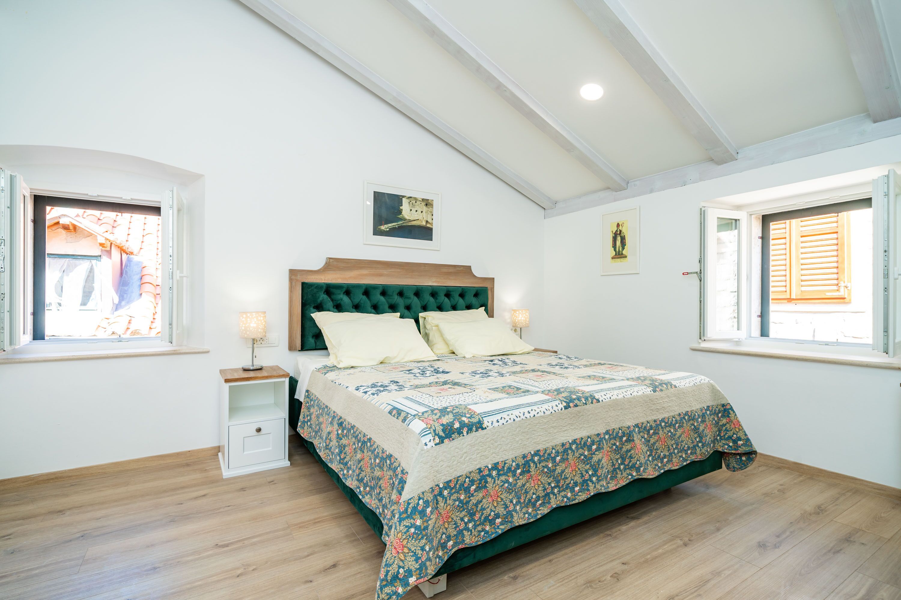 Un dormitorio con una cama de madera, una mesita de noche, un ventanal con contraventanas y un cuadro en la pared.