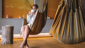 A person relaxing in a hammock in a room with wooden flooring, a wooden stool, and a wall with a sunset mural. - Haubers Naturresort (Oberstaufen)