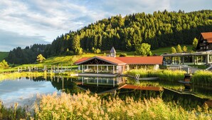 A lakeside lodge with a covered deck, a small tower, and a building with a red roof. - Haubers Naturresort (Oberstaufen)