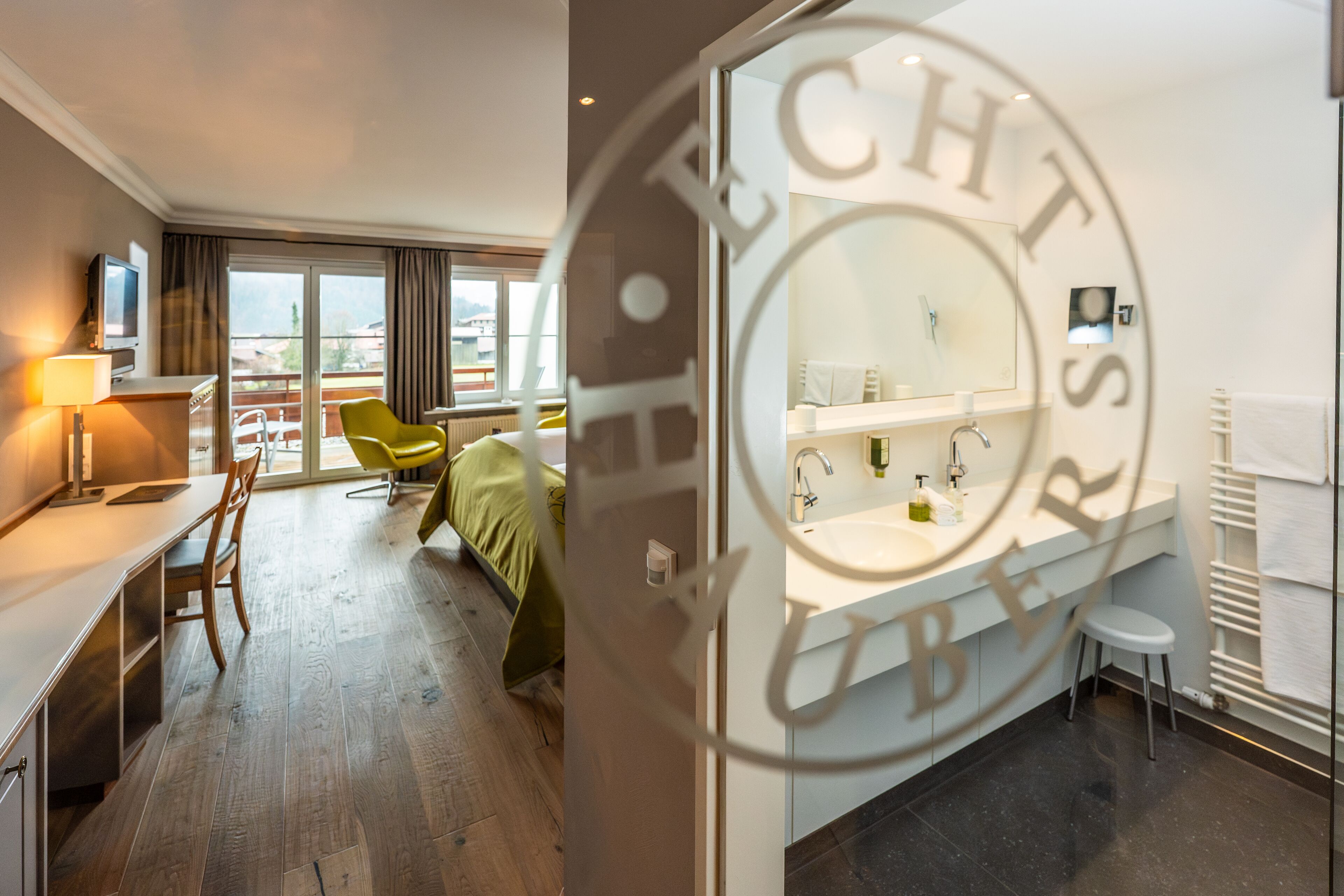 A hotel room with a bed, a desk, a chair, a TV, a window, and a bathroom with a sink and mirror.