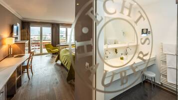 A hotel room with a bed, a desk, a chair, a TV, a window, and a bathroom with a sink and mirror.