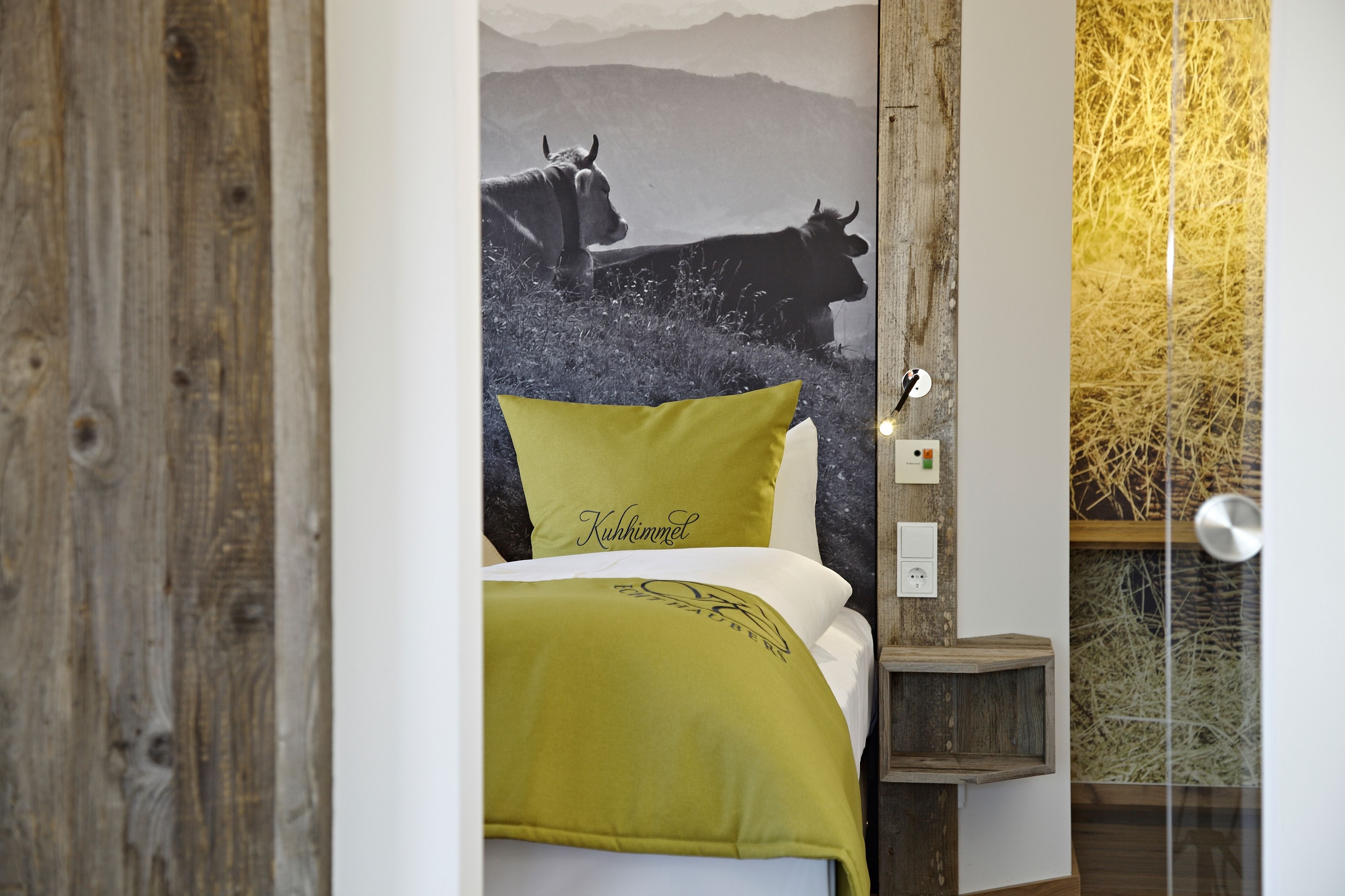 Ein Schlafzimmer mit einem Bett, einem Nachttisch, einer Wandlampe und einem Poster mit Kühen in den Bergen.
