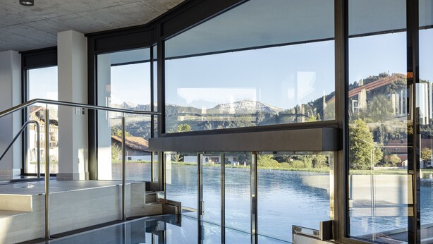 A modern indoor swimming pool with glass walls and a view of a lake and buildings. - Haubers Naturresort (Oberstaufen)