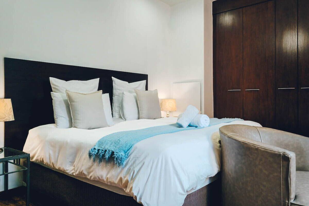A neatly made bed with a dark headboard, a blue throw blanket, and multiple pillows. There is a dark wooden wardrobe to the right and a small table with a lamp beside the bed.
