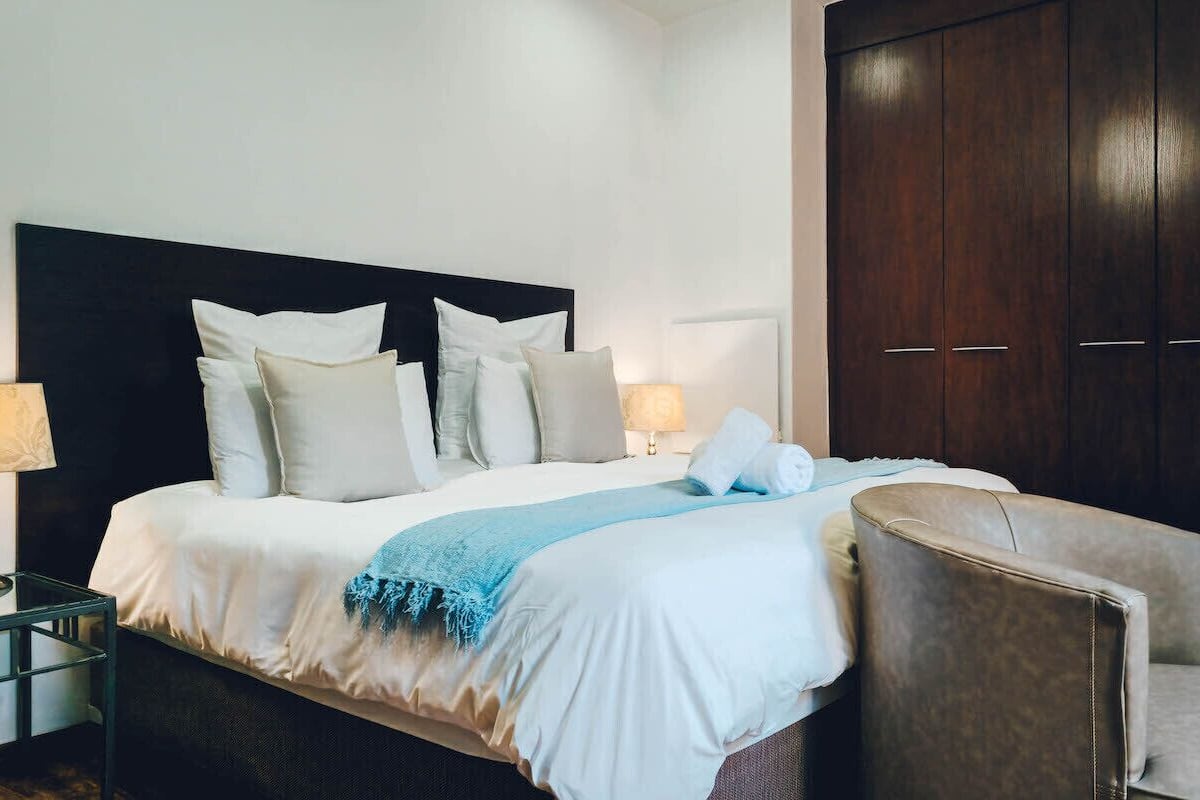 A neatly made bed with a dark headboard, a blue throw blanket, and multiple pillows. There is a dark wooden wardrobe to the right and a small table with a lamp beside the bed.