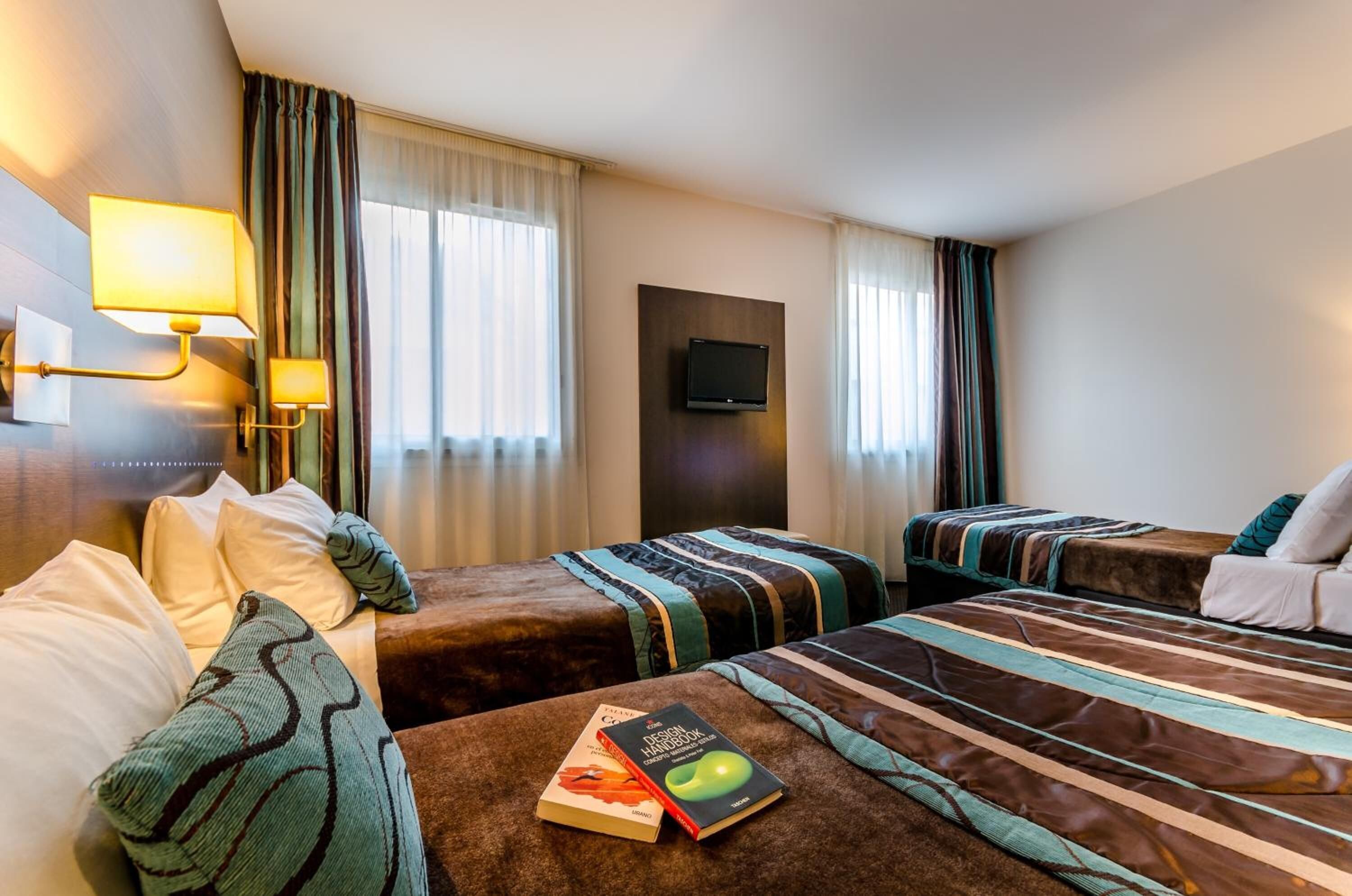 A hotel room with two single beds, a sofa, a flat-screen TV mounted on the wall, and a window with curtains.