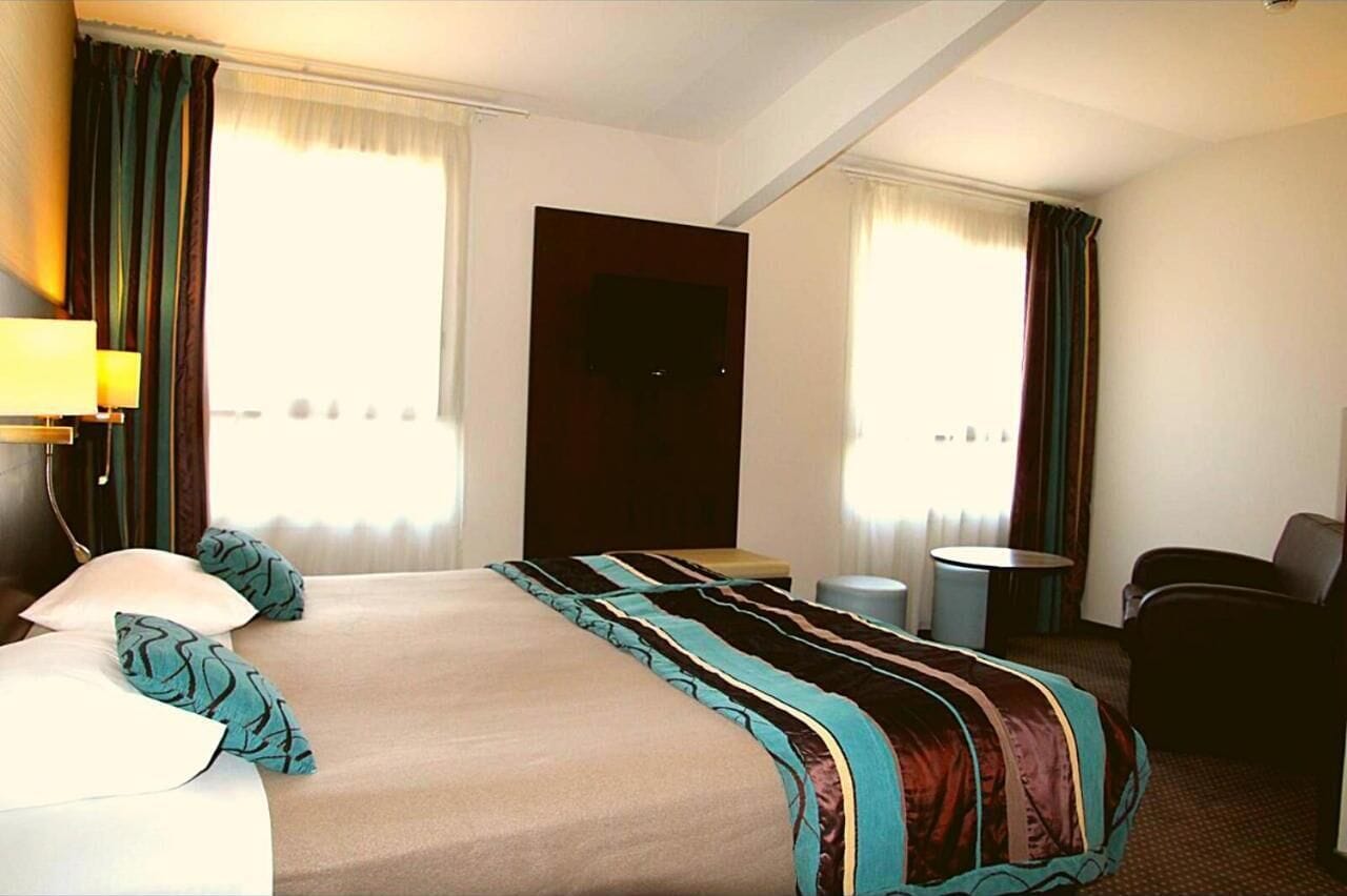 Habitación de hotel con una cama grande, un escritorio, una silla, un televisor y dos ventanas con cortinas.