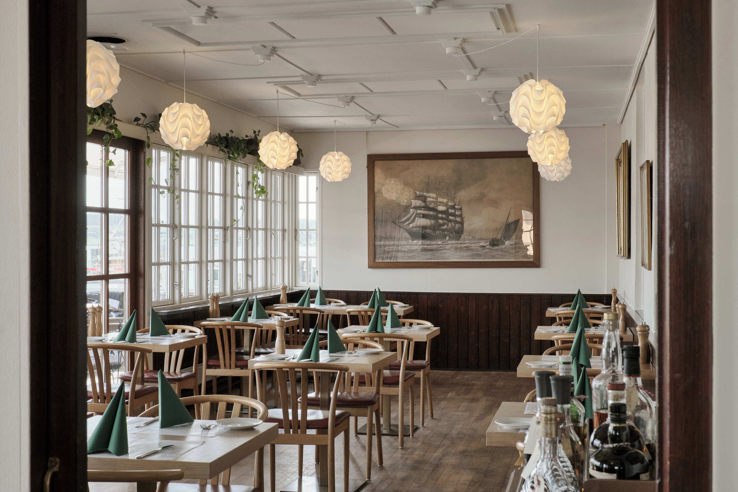 Un restaurante con mesas y sillas de madera, lámparas colgantes y un cuadro grande de un barco en la pared.