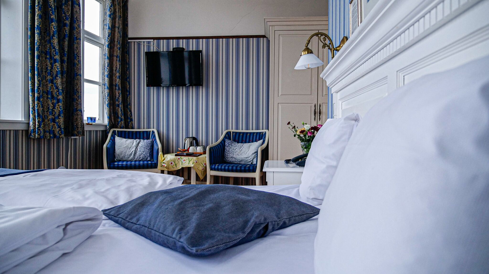 A hotel room with two beds, a TV, two armchairs, a small table with a coffee pot, and a vase with flowers.