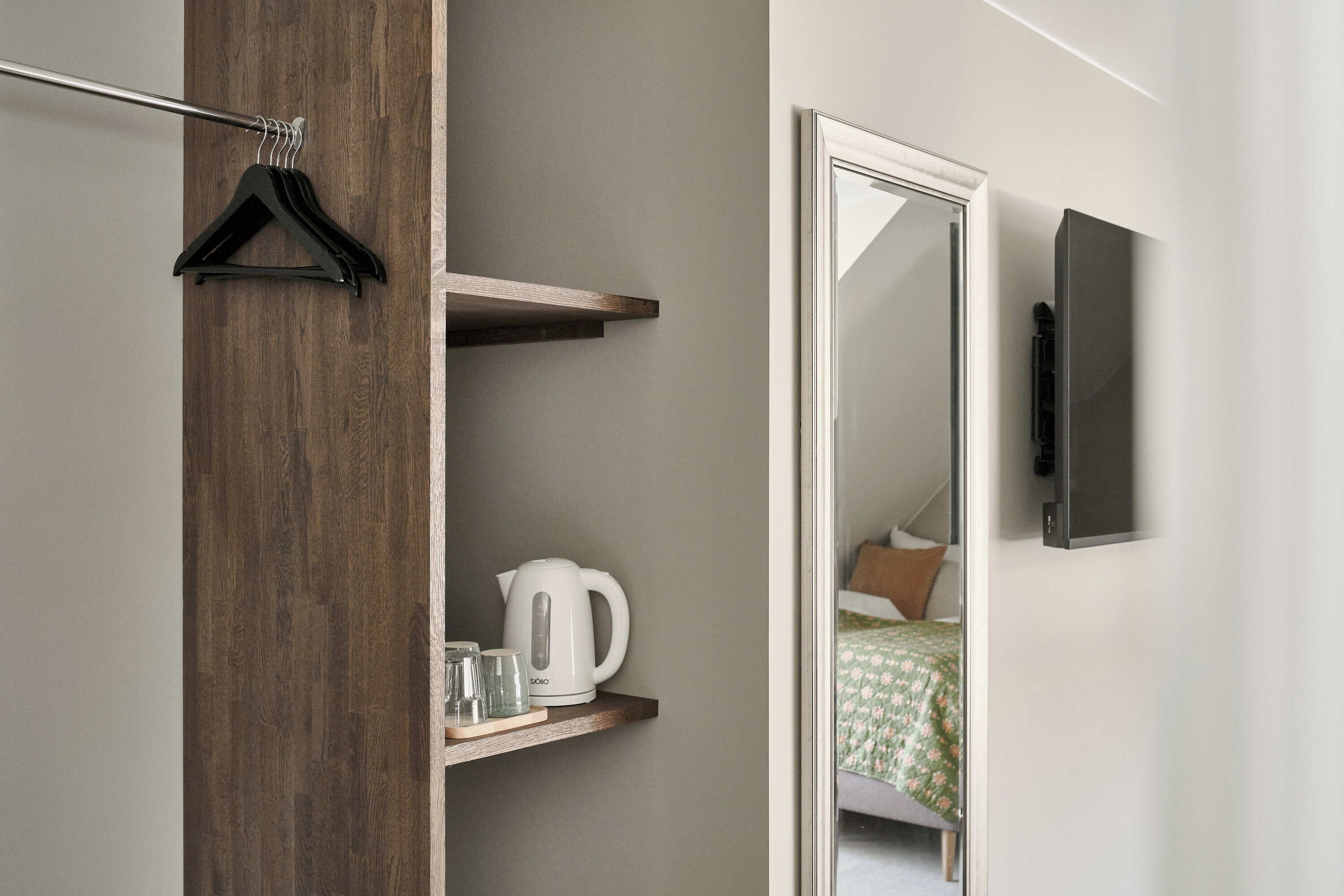 Una habitación de hotel moderna con un armario de madera, una tetera blanca y una pequeña estantería con vasos.