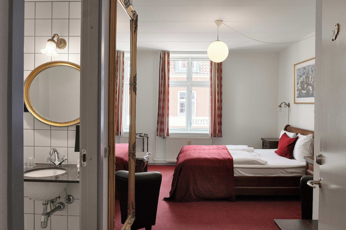A hotel room with a large mirror, a round sink, a bed with red bedding, and a window with curtains.