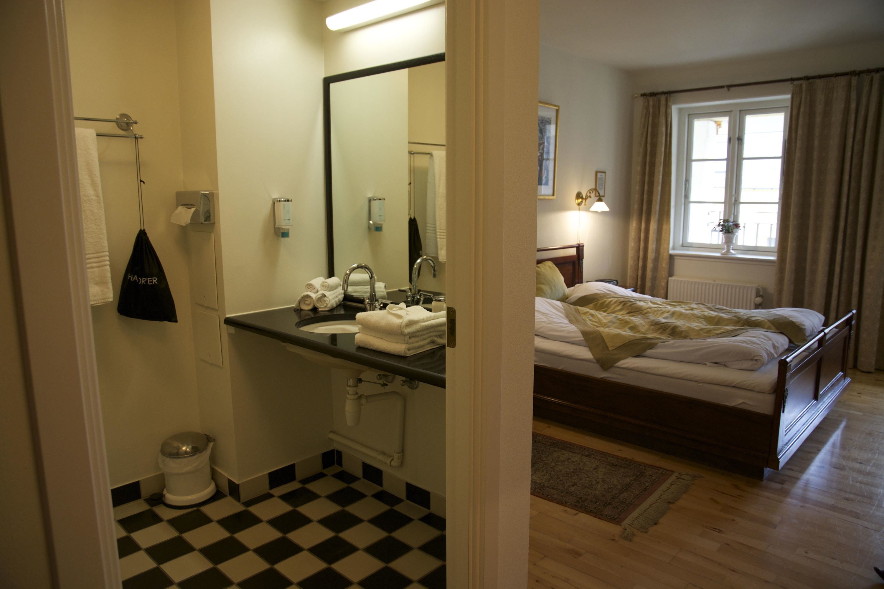 Una habitación de hotel con suelo a cuadros, una cama con edredón blanco, un baño con lavabo y espejo, y una ventana con cortinas.
