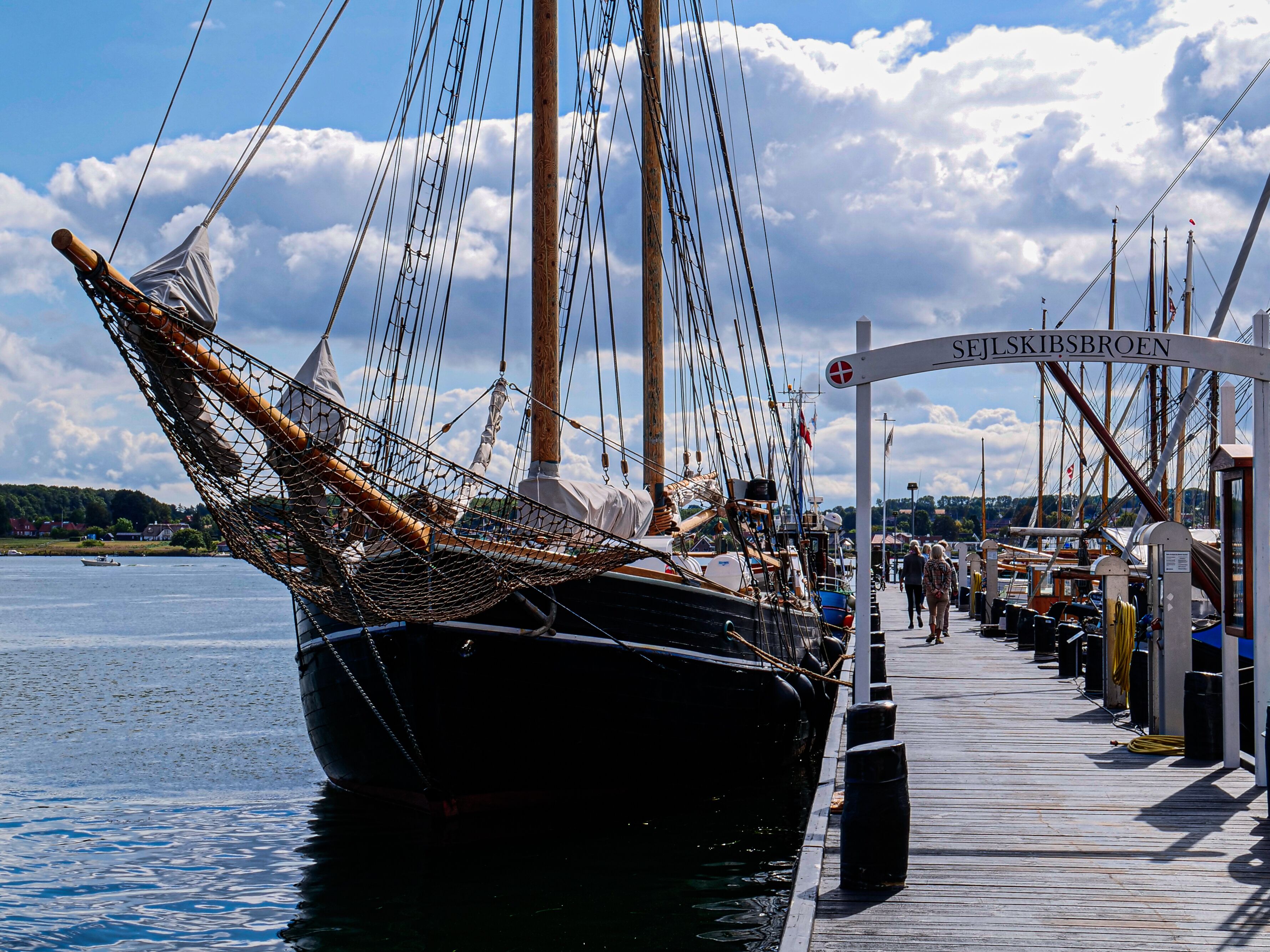 A wooden pier with a sailing ship docked, a sign reading "SEJLSKIBSBROEN", and a calm body of water in the background.