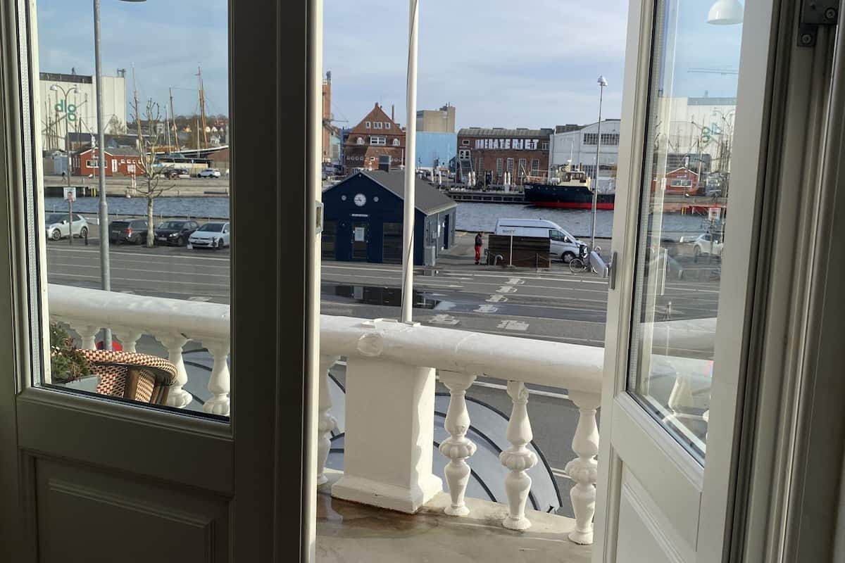 A view from a balcony with a railing, overlooking a waterfront area with buildings, parked cars, and a dock.
