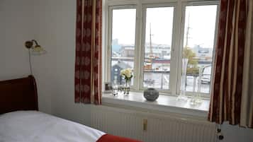 A bedroom with a bed, a window with curtains, a radiator, and a view of the city.