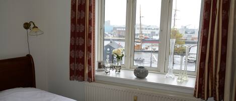 Un dormitorio con cama, ventana con cortinas, radiador y vistas a la ciudad.