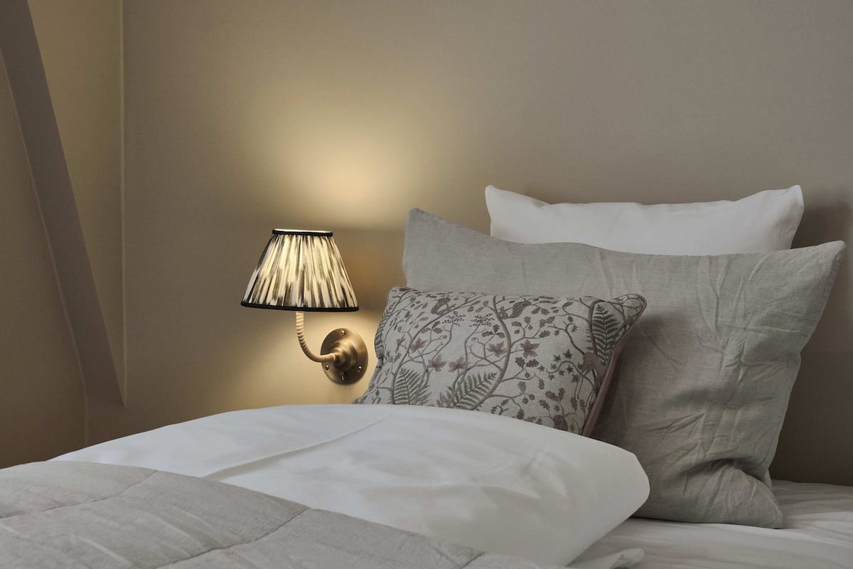 A neatly made bed with a white comforter and pillows, a wall-mounted lamp, and a textured wall.