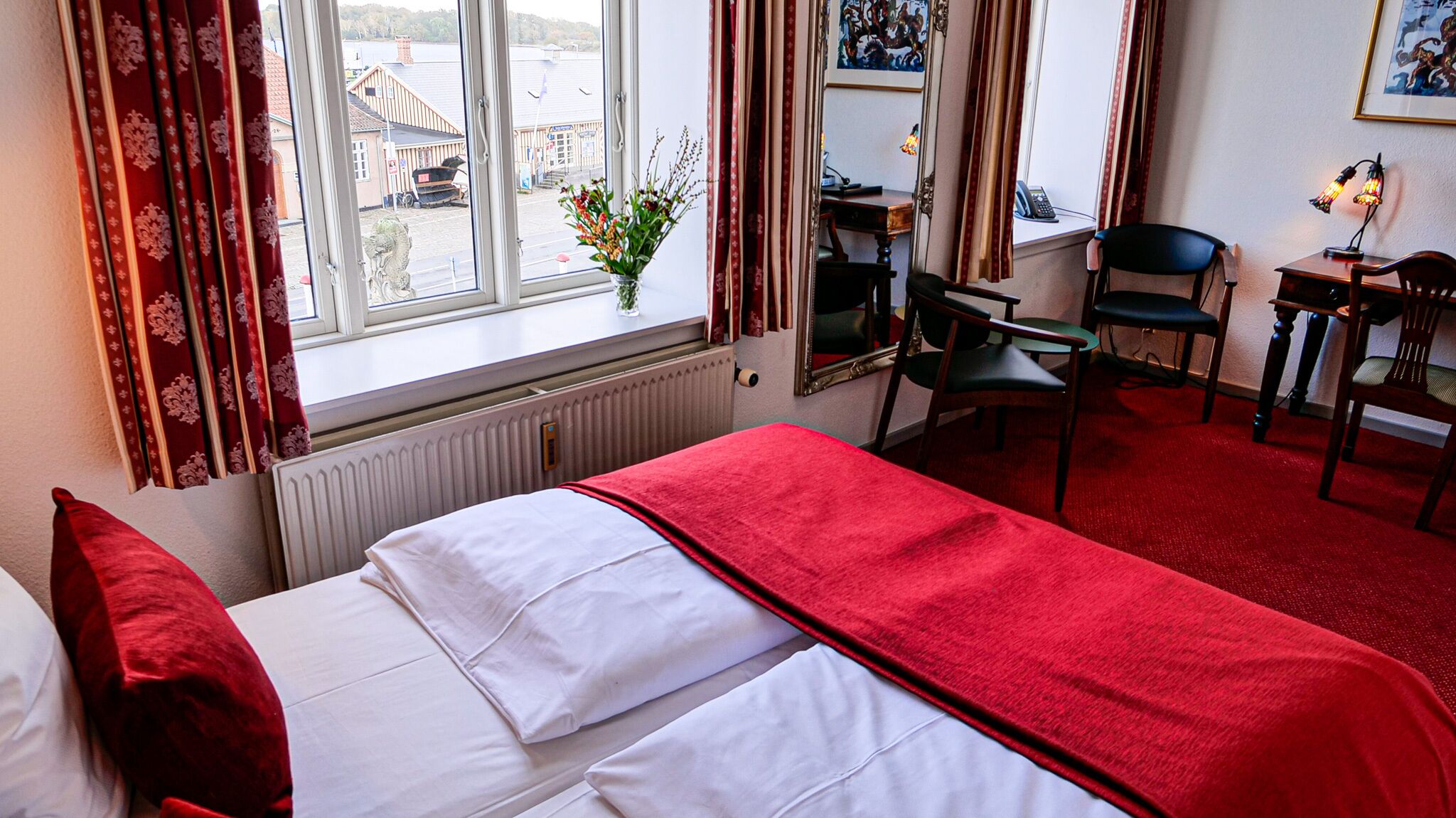 A hotel room with a bed, a desk, and a chair. There is a window with curtains, a radiator, and a plant on the windowsill.