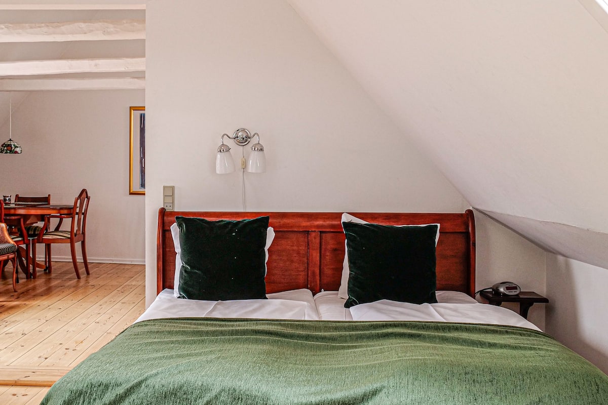 A bedroom with a wooden headboard, two green pillows, a green blanket, and a bedside table with a clock.