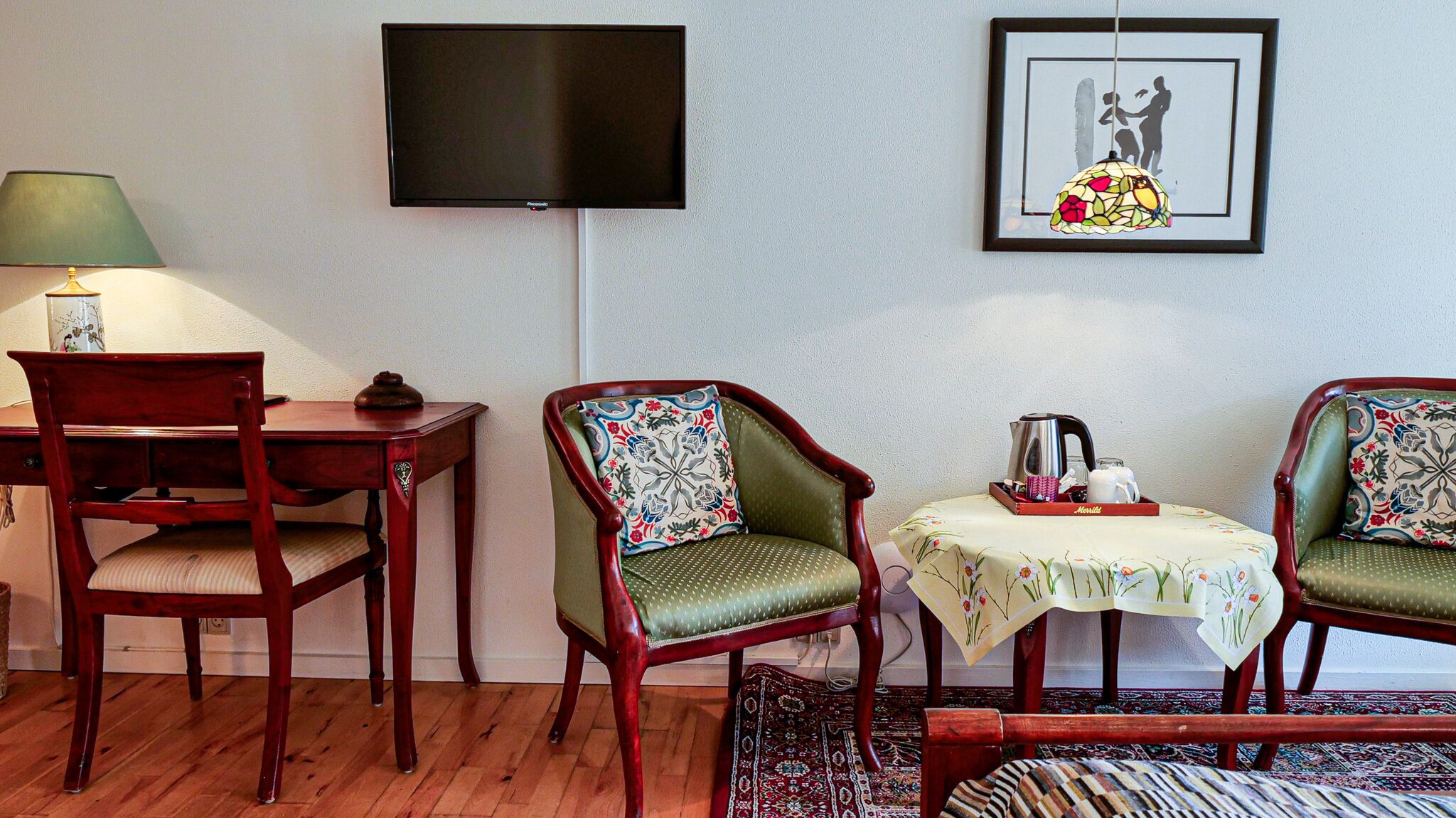 A room with a wooden desk, two upholstered chairs, a small table with a tray, a television mounted on the wall, and a framed picture.
