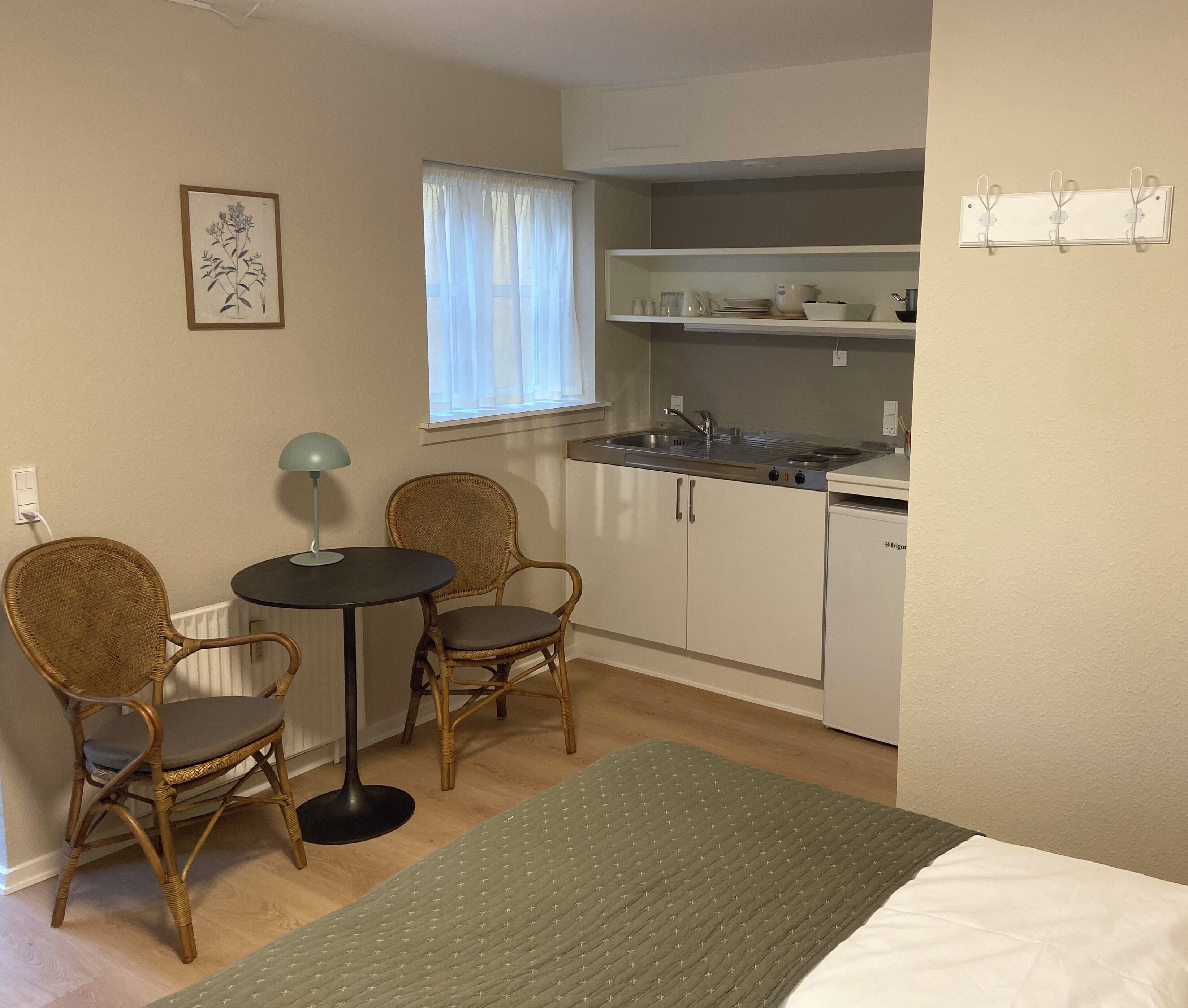 Une pièce de taille modeste comprenant un lit, une petite table, deux chaises en osier, une kitchenette avec un évier et une cuisinière, et une fenêtre ornée de rideaux légers.