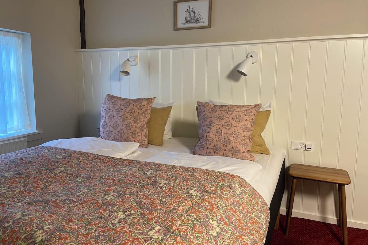 A neatly made bed with patterned bedding, a small wooden stool, and a framed picture on the wall.