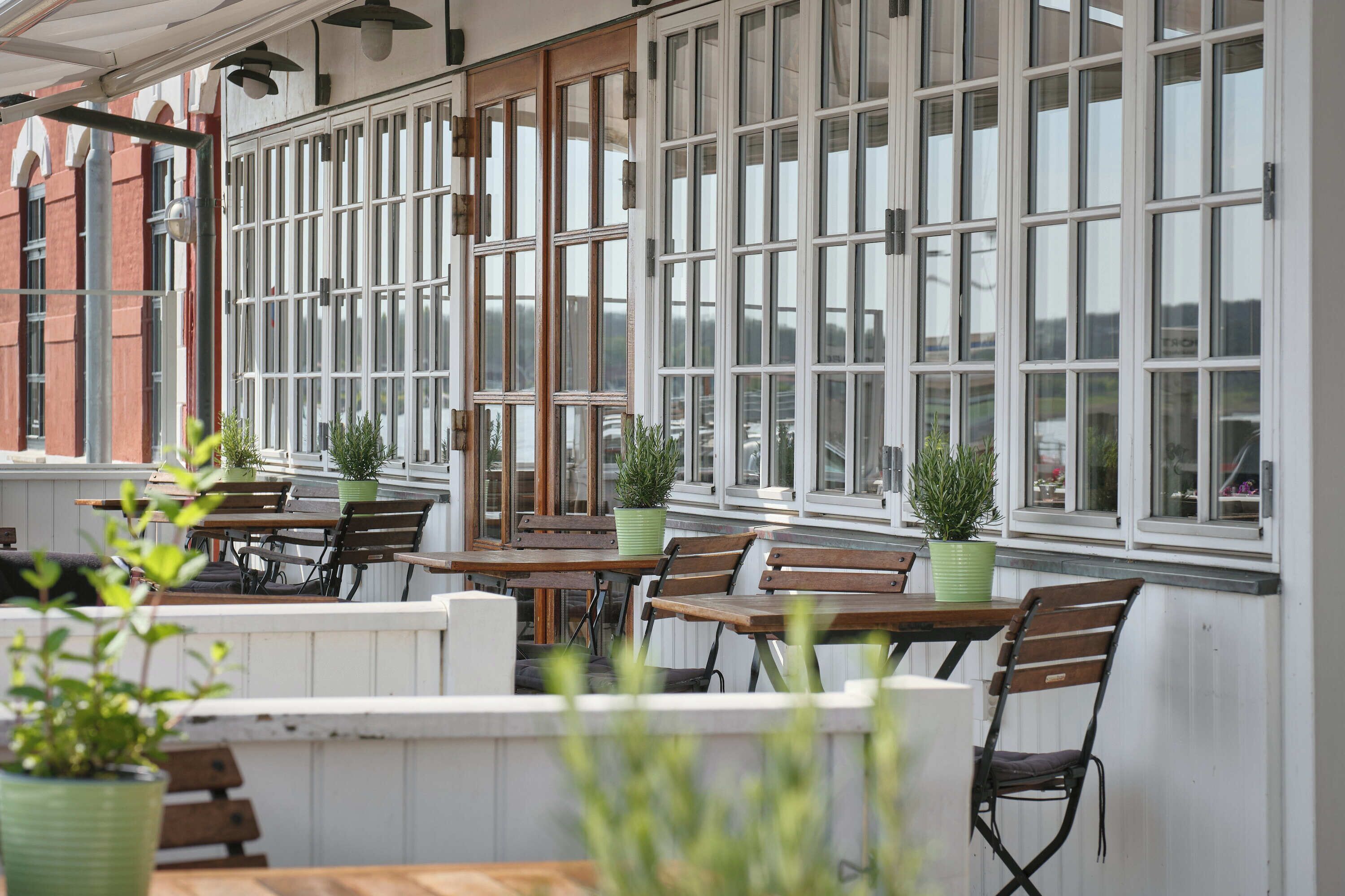 An outdoor seating area with wooden tables and chairs, potted plants, and large windows.