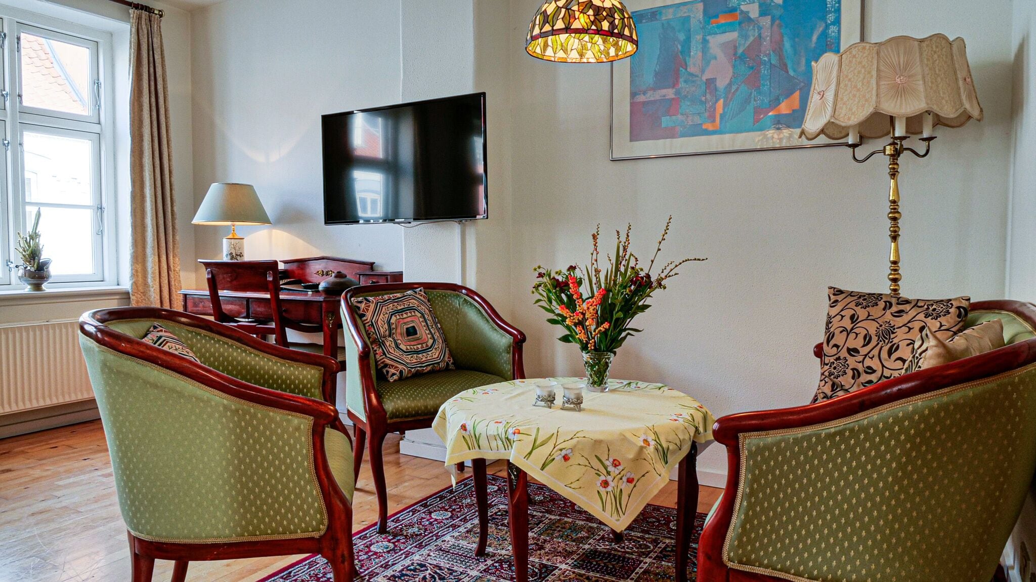 Una sala de estar con televisión, dos sillones, una mesita con mantel de flores, una lámpara y un cuadro en la pared.