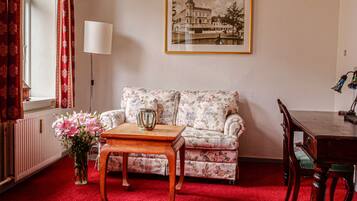 Una sala de estar con un sofá floral, una mesa de centro de madera, un jarrón con flores, una lámpara y un cuadro en la pared.