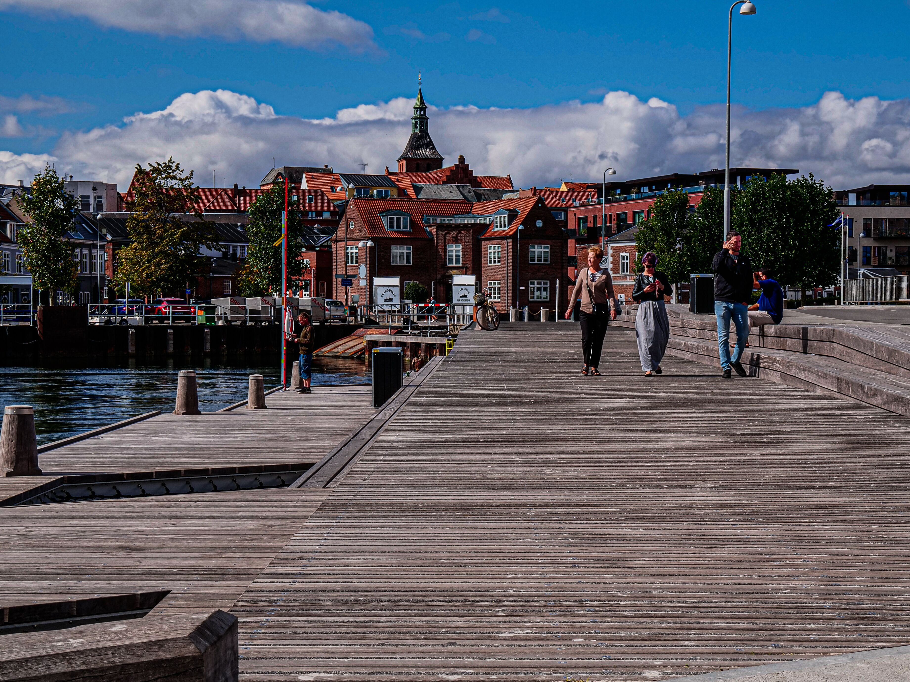 En træbro, der strækker sig ud i vandet, med bygninger i baggrunden.