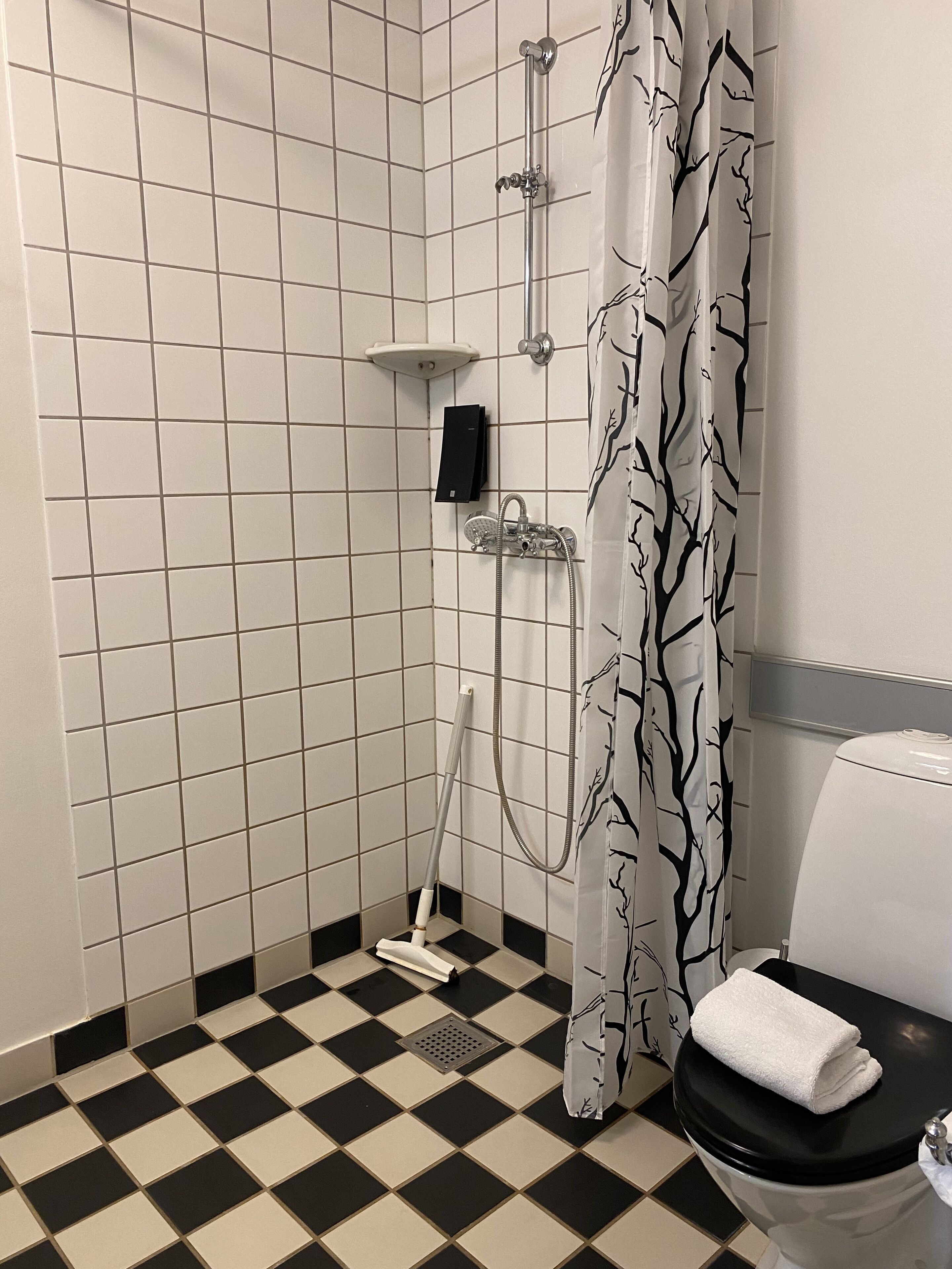 Casa de banho com chão xadrez, chuveiro com cortina estampada com padrão de árvore, sanita e chuveiro manual.
