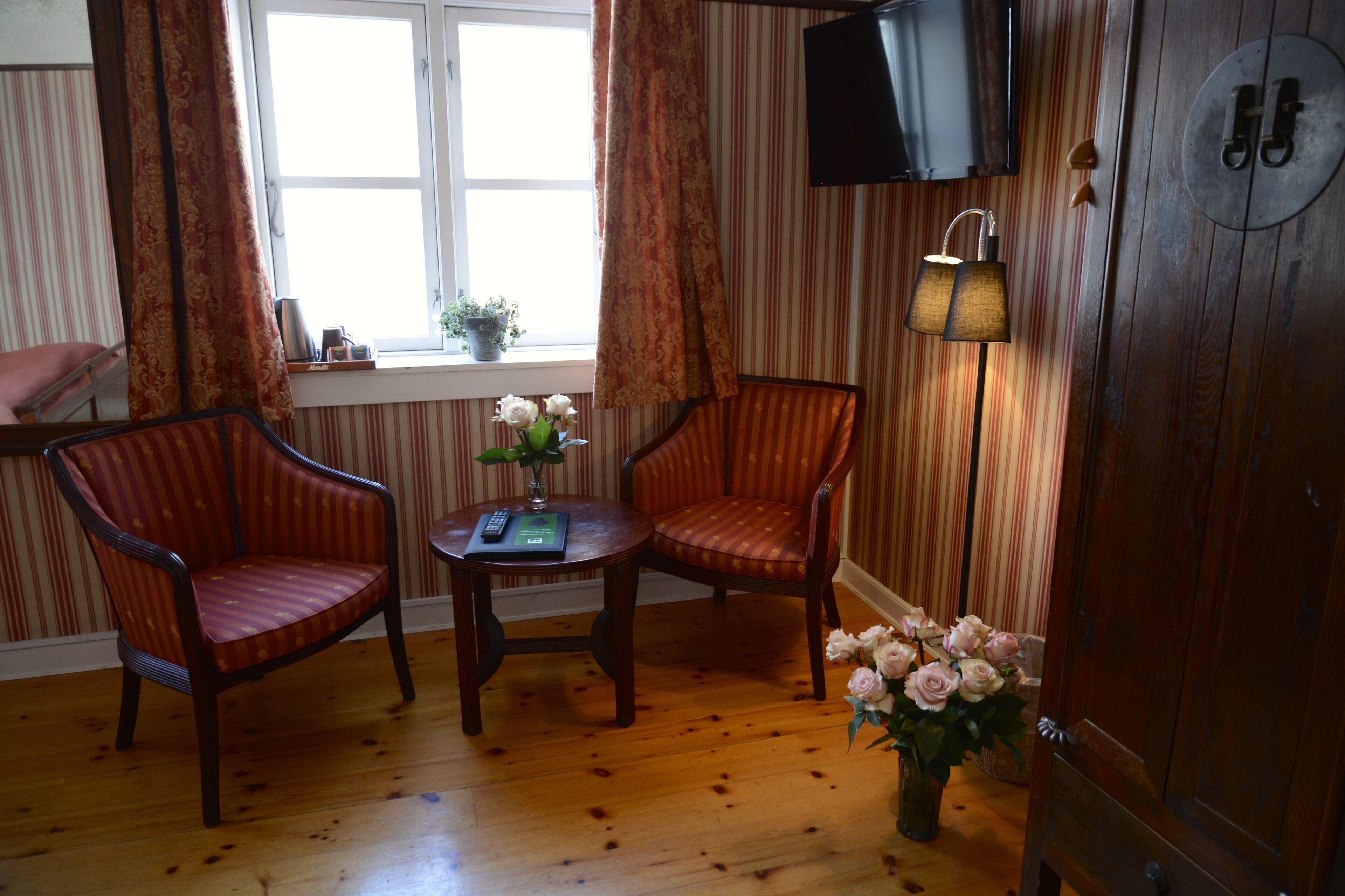 A room with two armchairs, a small table with a vase of flowers, a television mounted on the wall, and a window with curtains.