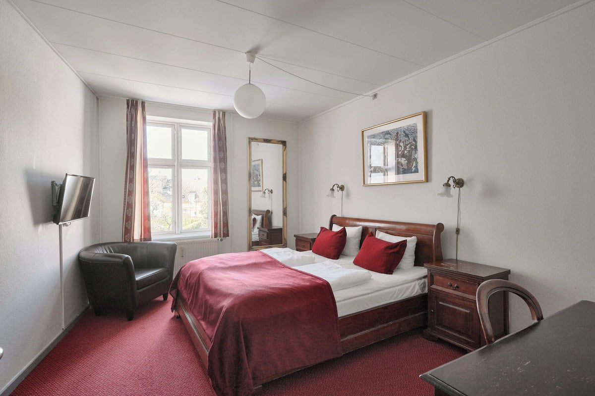 A bedroom with a bed, a chair, a desk, a TV, and a window with curtains.