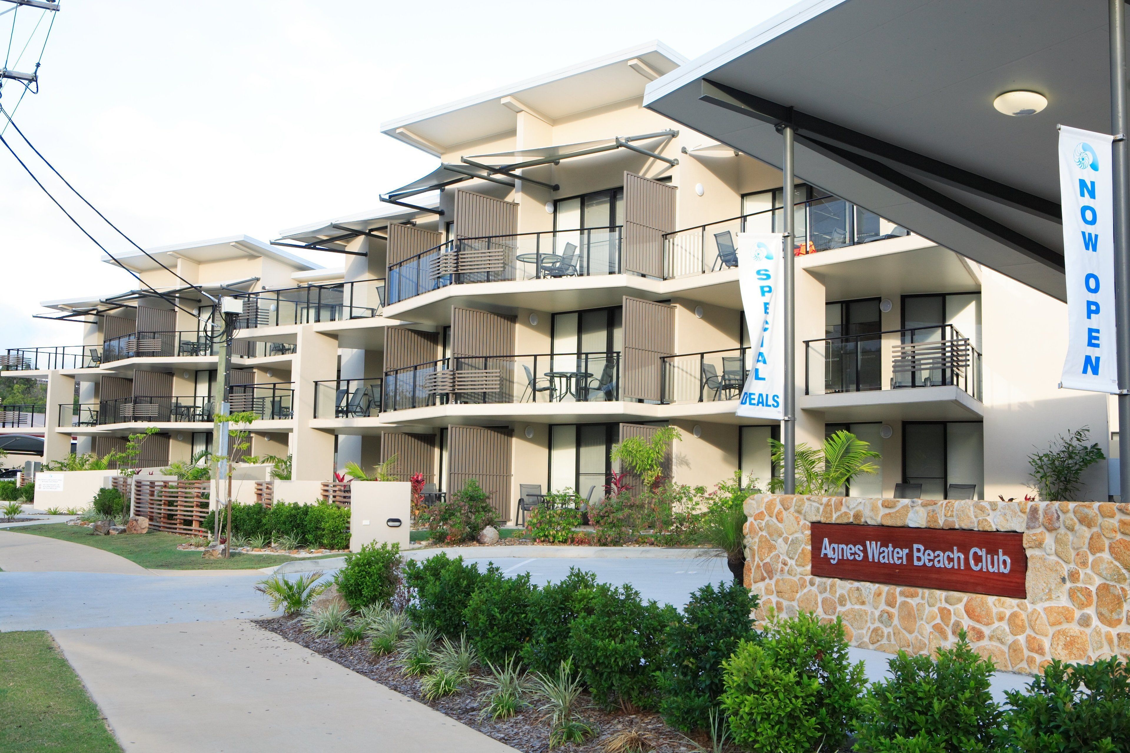 Un hotel de varios pisos con balcones, un cartel que indica 'NOW OPEN' y un letrero que dice 'Agnes Water Beach Club'.