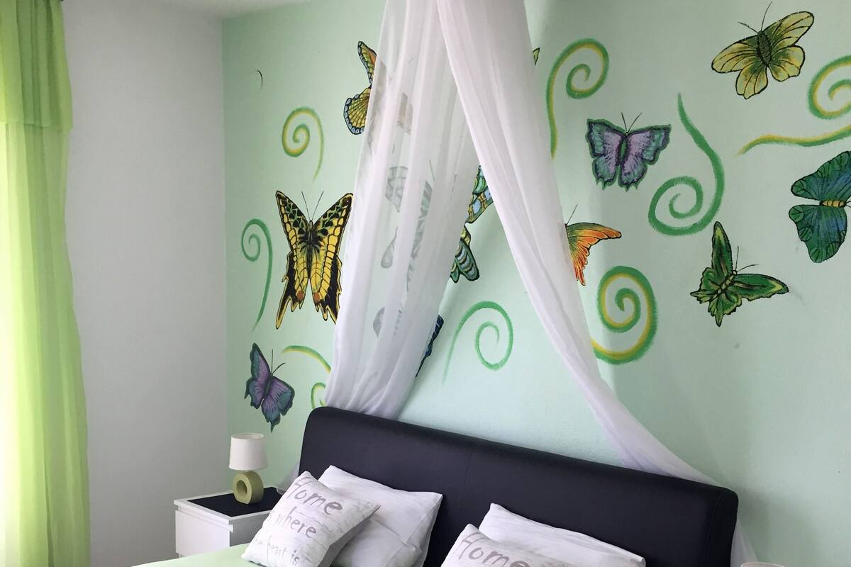 A bedroom with a bed, green curtains, butterfly wall art, and matching pillows.