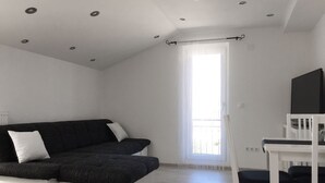 A modern, minimalist room with a sectional sofa, a white desk, and a large window with blinds. - Logos Guesthouse (Okrug)