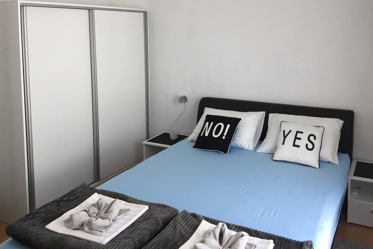 A neatly made bed with a blue mattress, two pillows with 'NO!' and 'YES' text, and a bedside table with a lamp.