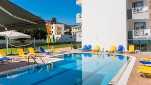 A swimming pool with lounge chairs and umbrellas, a building with balconies, and a clear blue sky. - Vagelis Comfort Apartments (Hersonissos)