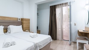 A hotel room with two beds, a wooden headboard, a desk, and a sliding glass door leading to a balcony. - Vagelis Comfort Apartments (Hersonissos)